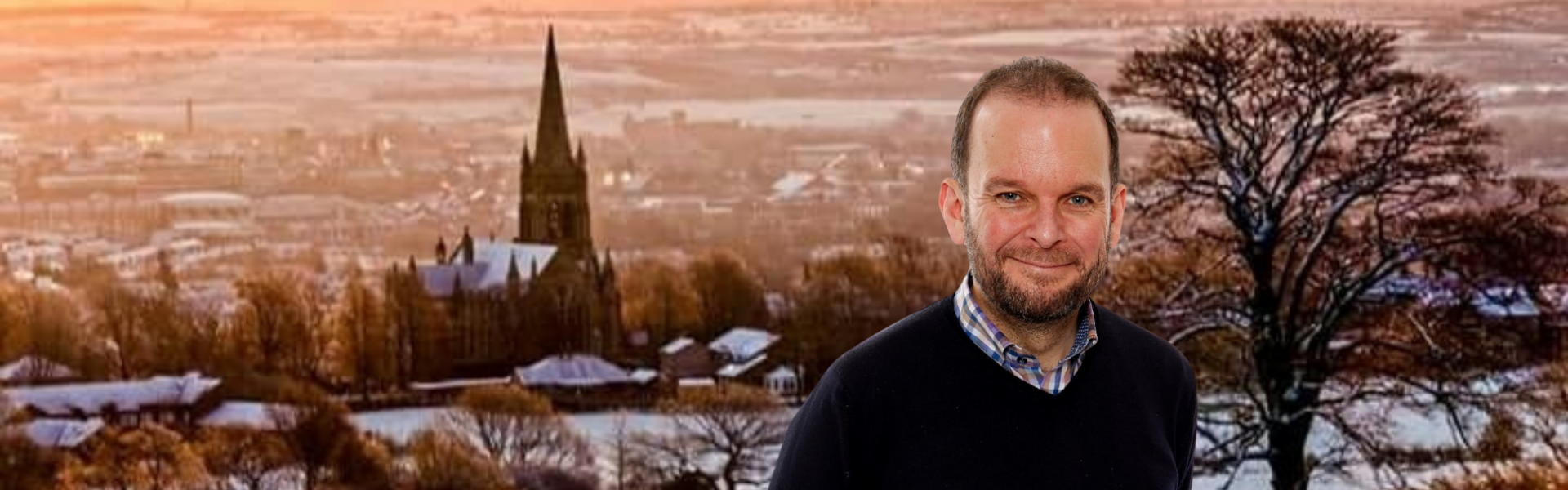  The image presents James Daly, Conservative Candidate before a snow-draped Walshaw backdrop. His attire is casual with a dark sweater over a checkered shirt. The winter scene behind him showcases a church spire amid the snow-blanketed village, under a warm-hued sky.