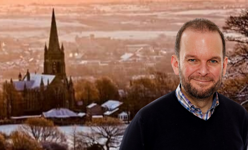  The image presents James Daly, Conservative Candidate before a snow-draped Walshaw backdrop. His attire is casual with a dark sweater over a checkered shirt. The winter scene behind him showcases a church spire amid the snow-blanketed village, under a warm-hued sky.
